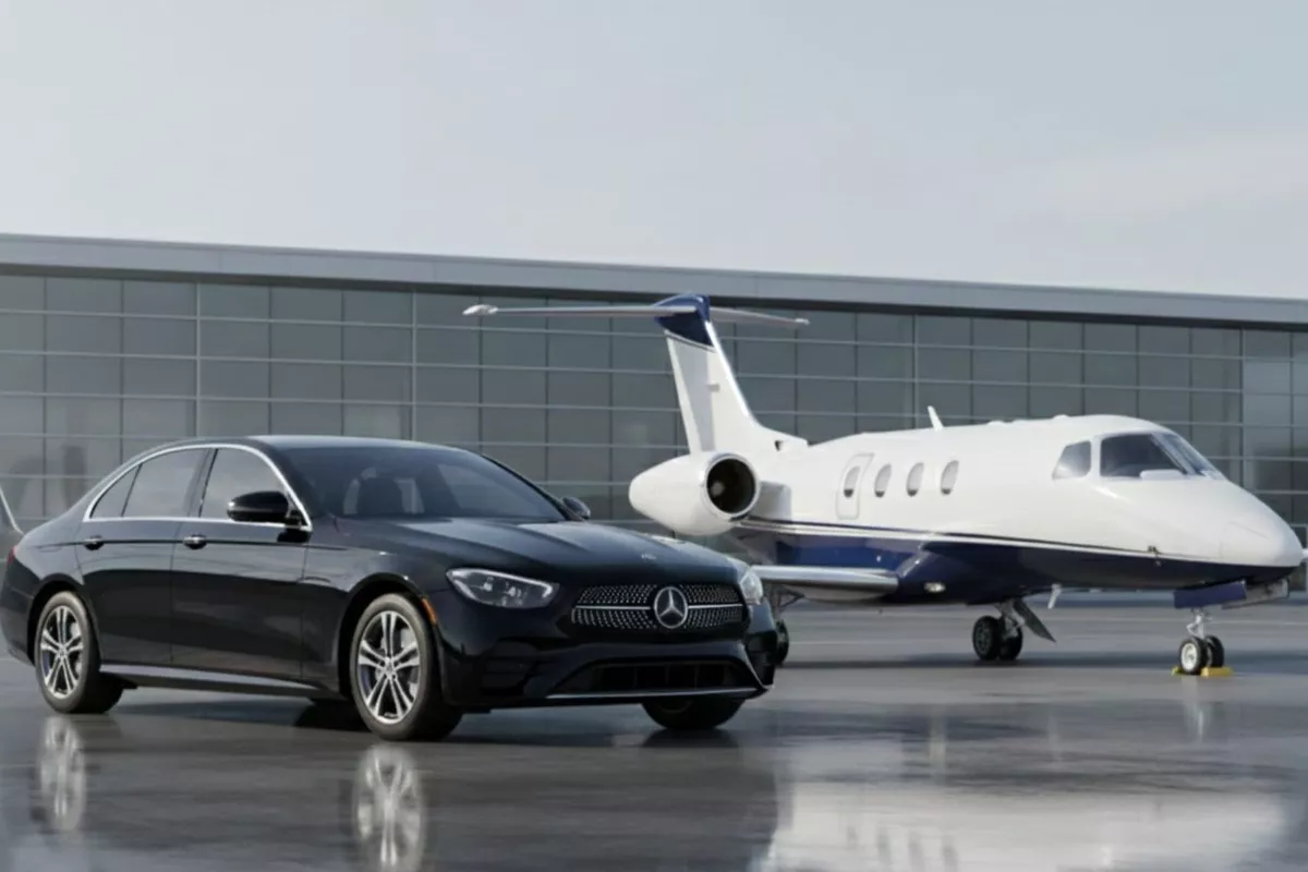 Berline Mercedes-Benz noire garée sur un tarmac d'aéroport à côté d'un jet privé blanc et bleu marine, devant un bâtiment moderne en verre et acier